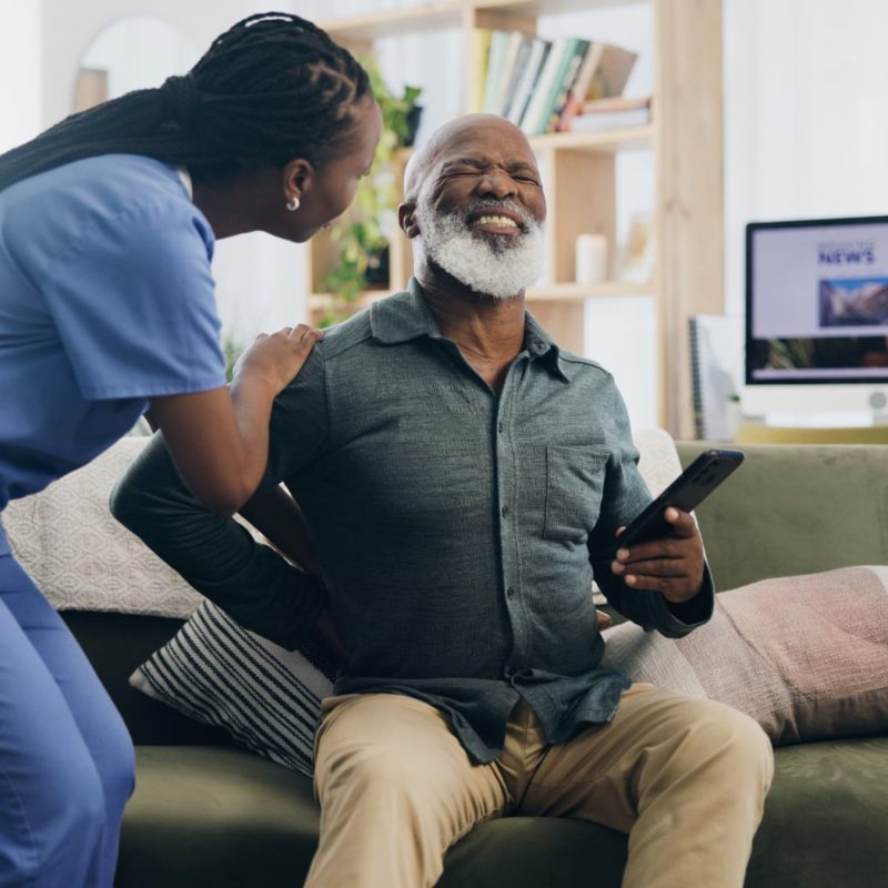 nurse-senior-man-back-pain-sofa-help-care-support-recovery-from-injury-retirement-black-people-physical-therapy-elderly-person-with-muscle-rehabilitation-watching-tv